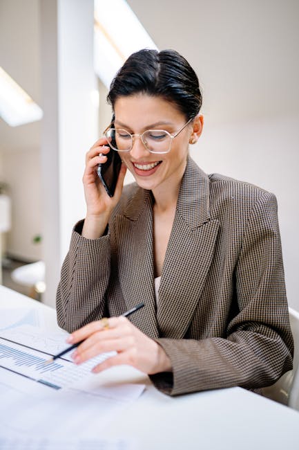 Confident businesswoman wearing glasses, talking on phone while reviewing documents in modern office setting.